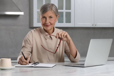 Güzel son sınıf öğrencisi, mutfaktaki beyaz mermer masada dizüstü bilgisayarın yanında notlar alıyor.