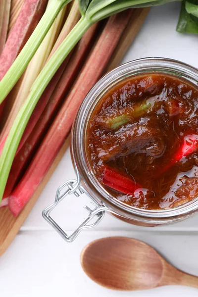 Jar of tasty rhubarb jam, stems and spoon on white wooden table, flat lay