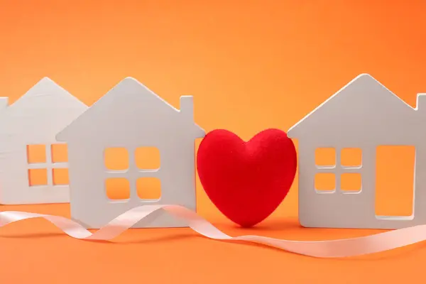 Long-distance relationship concept. Decorative heart between white house models and ribbon on orange background