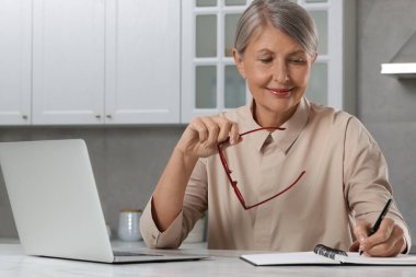 Güzel son sınıf öğrencisi, mutfaktaki beyaz mermer masada dizüstü bilgisayarın yanında notlar alıyor.