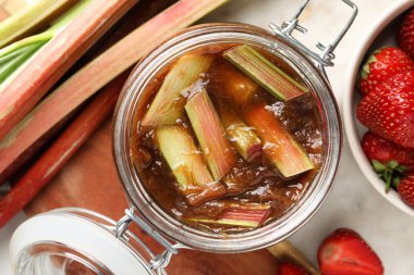 Lezzetli bir kavanoz ravent reçeli, taze saplar ve beyaz masa üzerinde çilek, düz uzanmış.