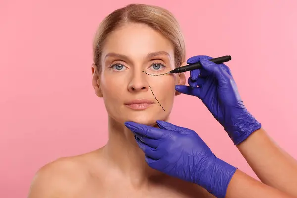 Woman preparing for cosmetic surgery, pink background. Doctor drawing markings on her face, closeup