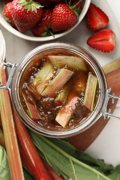 Jar of tasty rhubarb jam, fresh stems and strawberries on white table, flat lay