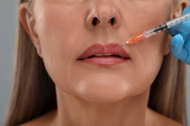 Doktor açık gri arka planda yaşlı kadına dudak iğnesi yapıyor, yakın plan. Estetik ameliyat