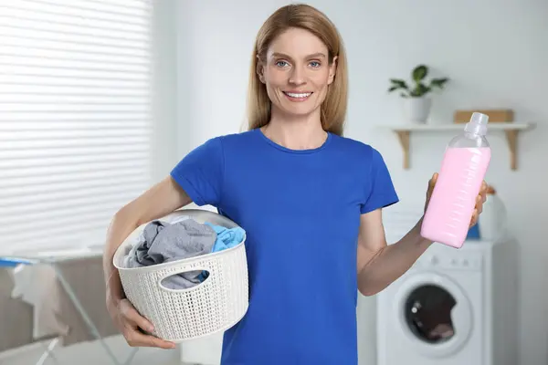 Woman holding fabric softener and basket with dirty clothes in bathroom