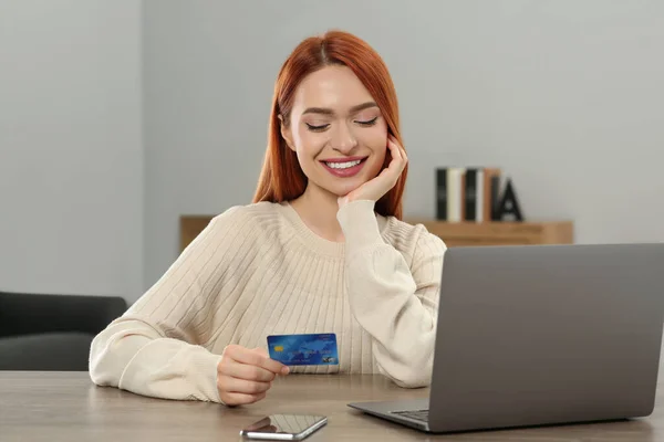 Happy woman with credit card using laptop for online shopping at wooden table in room