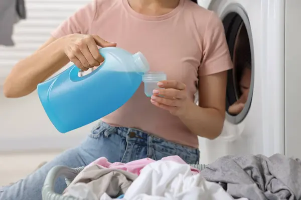 Woman pouring fabric softener from bottle into cap near washing machine in bathroom, closeup