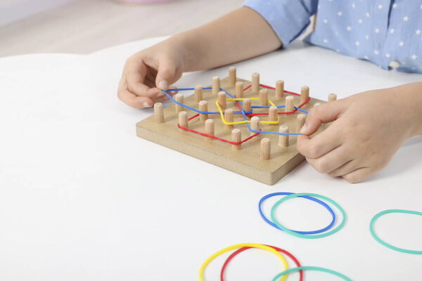 Motor skills development. Girl playing with geoboard and rubber bands at white table, closeup. Space for text