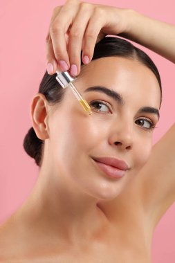 Güzel genç bir kadın pembe arka planda yüzüne serum sürüyor.