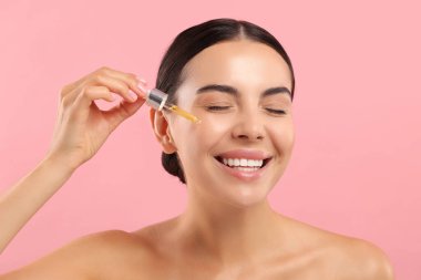 Güzel genç bir kadın pembe arka planda yüzüne serum sürüyor.