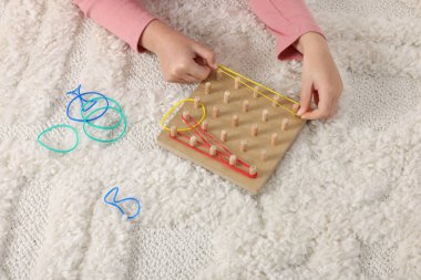 Motor skills development. Girl playing with geoboard and rubber bands on carpet, closeup clipart