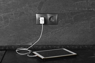 İçerideki koyu gri masada elektrik prizinden şarj olan tablet.