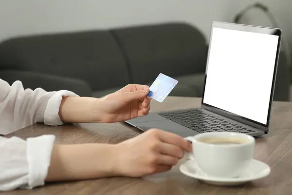 Woman with credit card and cup of coffee near laptop at wooden table indoors, closeup. Online shopping