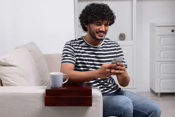 Happy man using smartphone on sofa with wooden armrest table at home