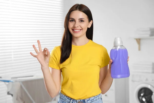 Woman holding fabric softener and showing OK gesture in bathroom