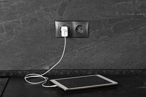 Tablet charging from electric socket on dark grey table indoors