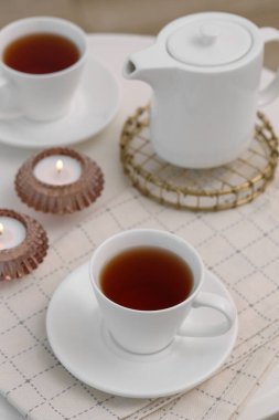Bir fincan çay, çaydanlık ve yanan mumlar beyaz masada.