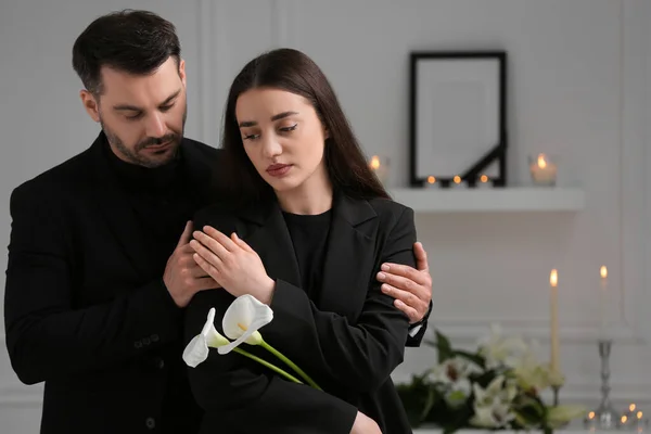 Sad couple with calla lily flowers mourning indoors, space for text. Funeral ceremony