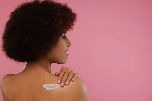 Beautiful young woman applying body cream onto back on pink background, space for text