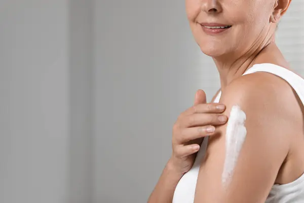 Happy woman applying body cream onto arm indoors, closeup. Space for text