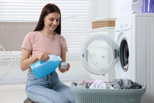 Woman pouring fabric softener from bottle into cap near washing machine in bathroom