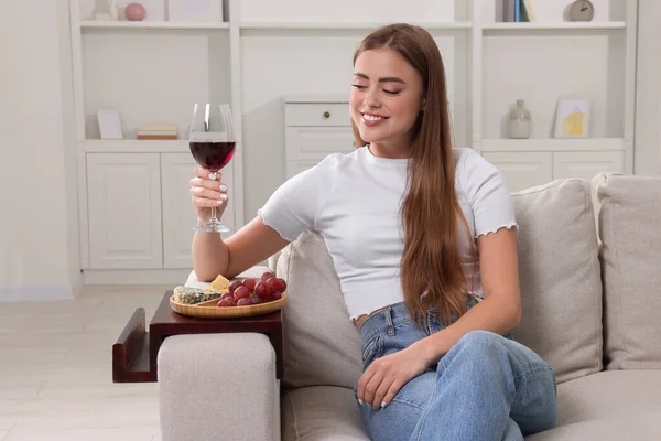 Happy woman holding glass of wine at home. Snacks on sofa armrest wooden table