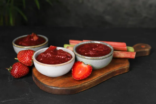 Tasty rhubarb jam in bowls, stems and strawberries on dark textured table