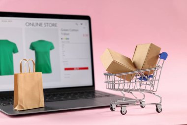 İnternetteki mağaza. Dizüstü bilgisayar, mini alışveriş arabası ve pembe arka planda alışverişler.