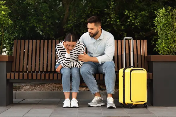 Long-distance relationship. Man calming his sad girlfriend on bench and suitcase outdoors