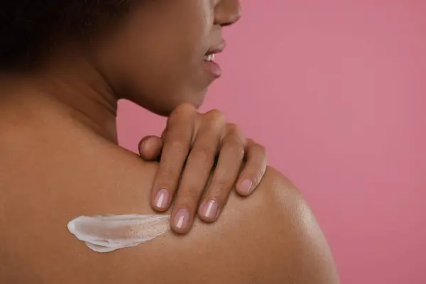 Young woman applying body cream onto back on pink background, closeup. Space for text