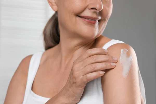 Happy woman applying body cream onto shoulder on light background, closeup