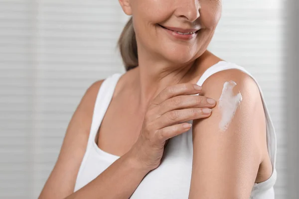 Happy woman applying body cream onto shoulder on light background, closeup