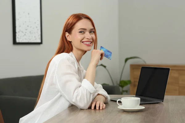 Happy woman with credit card near laptop at wooden table in room. Online shopping