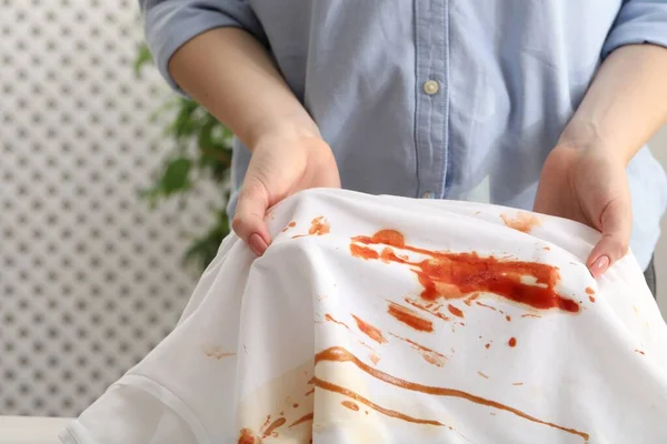 Woman holding shirt with stain of sauce on blurred background, closeup