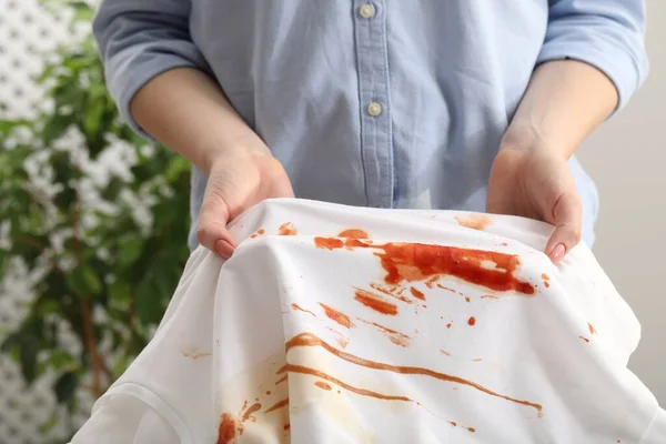 Woman holding shirt with stain of sauce on blurred background, closeup