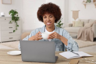 Laptop kullanan ve odasındaki ahşap masada kahve içen genç bir kadın.