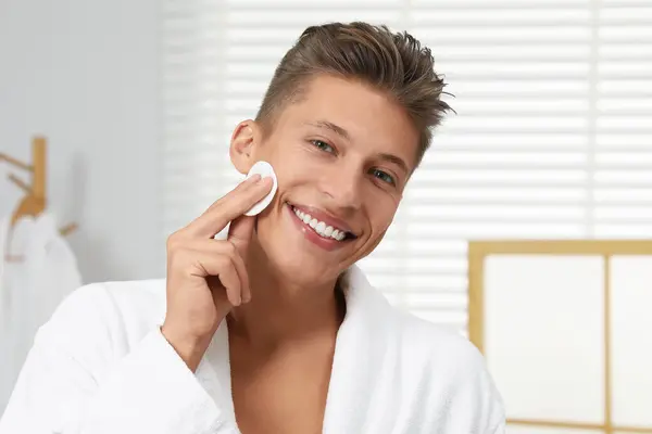 Handsome young man cleaning face with cotton pad in bathroom
