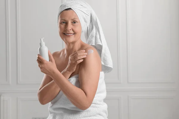 Happy woman applying body cream onto shoulder near white wall. Space for text