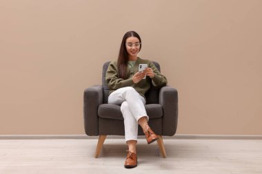 Akıllı telefonlu güzel bir kadın bej duvarın yanındaki koltukta oturuyor.