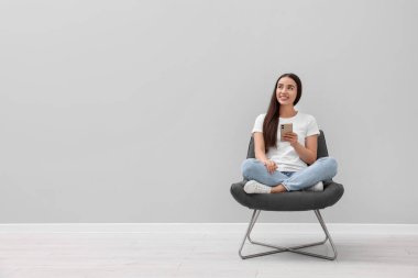 Akıllı telefonlu güzel bir kadın açık gri duvarın yanındaki koltukta oturuyor.