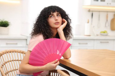 Mutfakta kendini serinletmek için pembe yelpaze sallayan genç bir kadın.