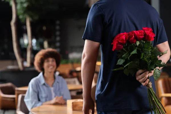 International dating. Man hiding bouquet of roses for his beloved woman in restaurant, closeup