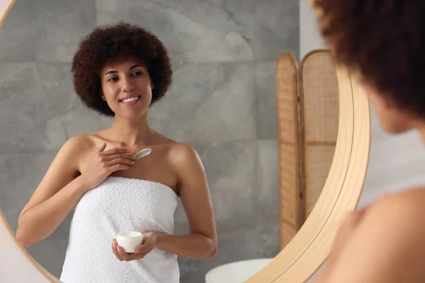 Beautiful young woman applying cream onto body in bathroom