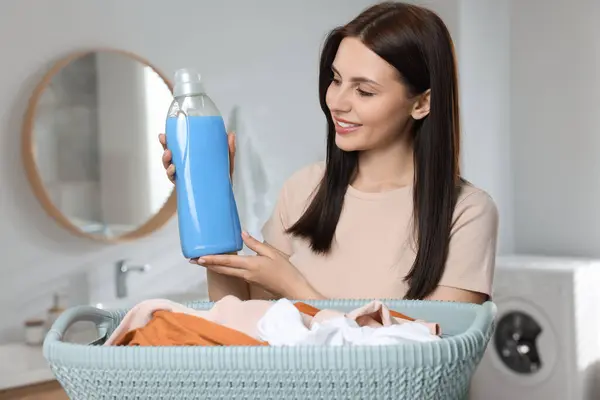 Woman holding fabric softener near basket with dirty clothes in bathroom