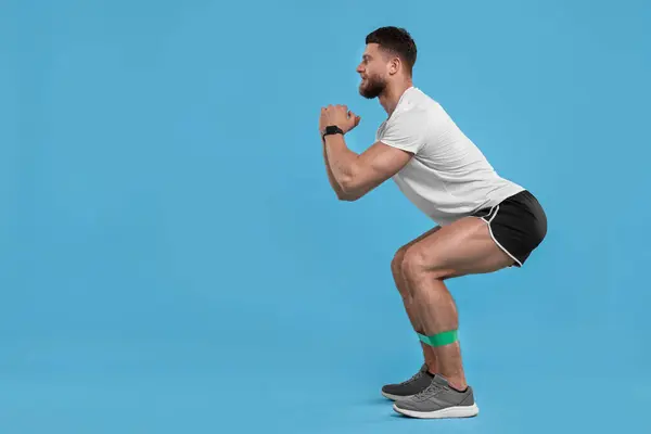 Young man exercising with elastic resistance band on light blue background. Space for text
