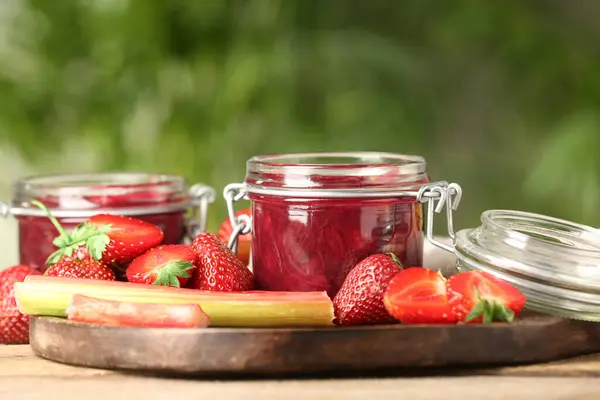 Tasty rhubarb jam in jars, stems and strawberries on wooden table against blurred background