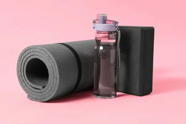 Grey exercise mat, yoga block and bottle of water on pink background