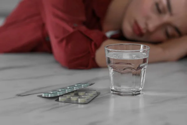 Woman sleeping at white marble table, focus on blisters with antidepressant pills and glass of water