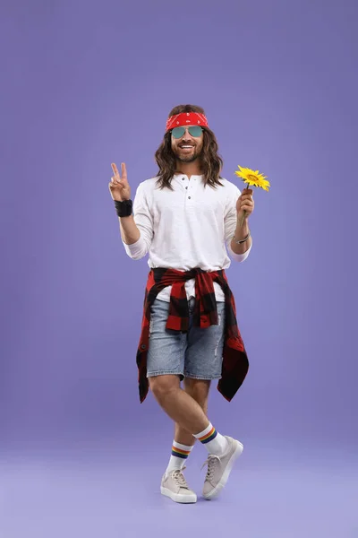 Stylish hippie man with sunflower showing V-sign on violet background