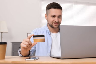 Odanın ahşap masasından online alışveriş için laptop kullanan, kredi kartı olan yakışıklı bir adam.